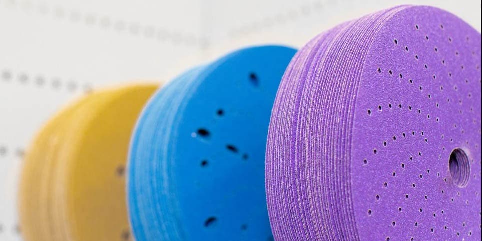 Rollos y discos abrasivos de diferentes colores y tamaños organizados sobre una mesa de trabajo.
