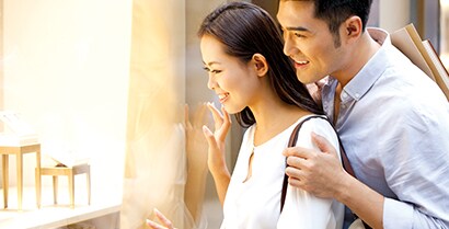 An Asian couple looks through a brightly lit window of a furniture store.
