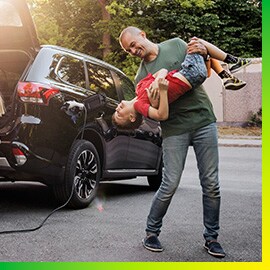 Man playing with son by a black car charing in driveway