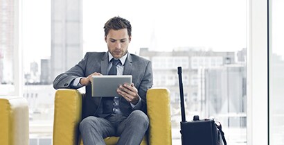 A professional man in a hotel lobby uses a tablet without worrying about glare on his screen from the wall of windows behind him.
