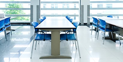 Sunlight shines through a serene scene of long study tables with blue chairs, providing a safe haven for students.
