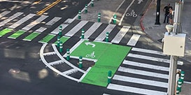 Urban intersection with crosswalks, protected bike lanes and two pedestrians on sidewalk.
