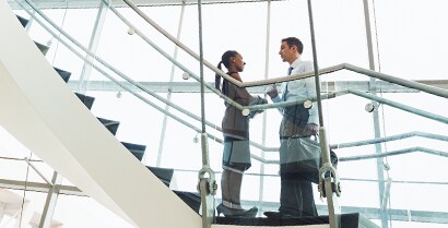A professionally dressed man and woman stop for a conversation on a stairway that is surrounded by floor-to-ceiling windows.
